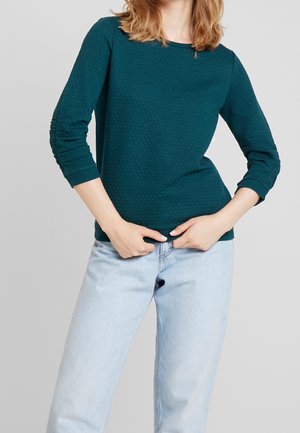 Vrouw die een tealkleurige top met textuur en lange mouwen en lichtblauwe spijkerbroek draagt, waarbij ze met beide handen de zoom van haar top rechtzet.