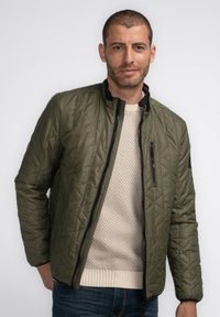 Grüne Steppjacke mit Stehkragen, ausgestattet mit einer seitlichen Reißverschlusstasche und schwarzen Akzenten. Getragen über einem cremfarbenen Strickpullover und blauen Jeans.