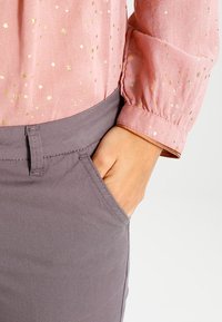 Rosa blus med gyllene stjärnmönster, rynkade ärmar, parat med grå byxor som har sidfickor och en slät textur.