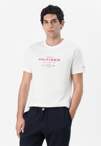 Bijela pamučna majica s crvenim logotipom "TOMMY HILFIGER" i grafikom. Kratki rukavi, okrugli izrez, u kombinaciji s tamnoplavim hlačama. Jednostavan dizajn.