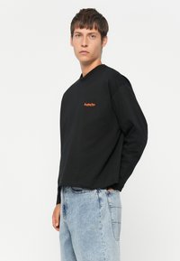Young man wearing a black long-sleeve shirt with orange Replay Inc. logo and light blue jeans, standing against a plain white background.