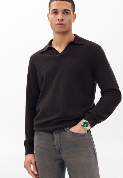 Pulover maro închis cu mâneci lungi și decolteu în V, realizat dintr-un material moale; purtat cu blugi albastru deschis, completat de un ceas argintiu cu cadran verde.