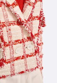 Gilet à carreaux rouge et blanc avec des bords effilochés, fait en tissu texturé. Il présente deux boutons blancs à l'avant, mettant en valeur des détails de design superposés.