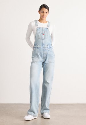 Lichtblauwe denim overalls met verstelbare schouderbanden, een voorzak en een wijd ontwerp. Gepaard met een witte langemouwen shirt en witte schoenen.