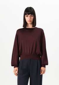 Vrouw met steil zwart haar, gekleed in een bordeauxrode blouse met lange mouwen en marineblauwe hoge taille broek, staande tegen een witte achtergrond.