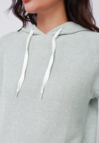 Lichtgroene hoodie met een gestructureerd, geweven patroon. Beschikt over een kraaglijn en witte trekkoorden. Close-up weergave van het schoudergebied.
