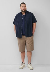 Marineblauwe korte mouwen overhemd over een blauwe t-shirt, gecombineerd met beige korte broeken en witte sneakers. Glad materiaal, casual pasvorm, minimale accenten.