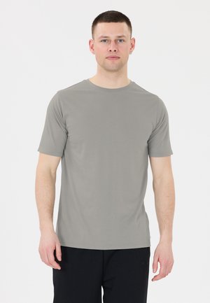 Junger Mann mit kurzen Haaren, der ein schlichtes graues kurzärmeliges T-Shirt und schwarze Hosen trägt, steht vor einem weißen Hintergrund.