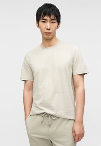 Ανοιχτό μπεζ μπλουζάκι με διακριτικό ολόκληρο μοτίβο. Κοντά μανίκια, στρογγυλή λαιμόκοψη, κατασκευασμένο από απαλό ύφασμα. Συνδυάζεται με μπεζ παντελόνι με κορδόνι στη μέση.