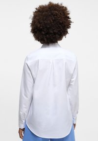 Camicia bianca a maniche lunghe con colletto button-down, presenta una piega sul retro e polsini con bottoni, realizzata in tessuto morbido.