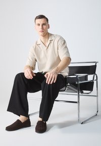 Beige Kurzarmhemd, schwarze weite Hose und braune gewebte Slipper. Sitzt auf einem Stuhl mit verchromtem Gestell und schwarzem Sitz. Minimalistischer Hintergrund.