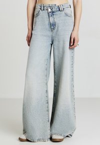 Jeans a gamba larga in denim lavato chiaro, con vita alta, chiusura frontale a bottone e classico design a cinque tasche. Texture morbida e sbiadita.