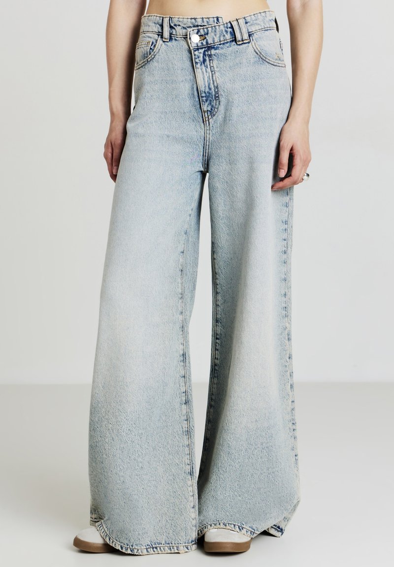 Jeans a gamba larga in denim lavato chiaro, con vita alta, chiusura frontale a bottone e classico design a cinque tasche. Texture morbida e sbiadita.