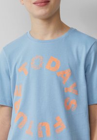 Helles blaues T-Shirt aus weichster Baumwolle, mit einem runden Grafikdesign und dem Text „DIE ZUKUNFT VON HEUTE“ in fettgedruckten orangefarbenen Buchstaben.