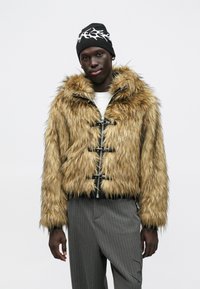 Casaco de pelúcia sintética castanho claro com detalhes a preto, com capuz, fechos de correr prateados e fechos de correr em alavanca. Usado com calças cinzentas aos risquinhos.