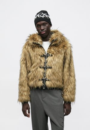 Kunstpelzjacke in Hellbraun mit schwarzen Akzenten, mit Kapuze, silbernen Reißverschlüssen und Knebelverschlüssen. Kombiniert mit grauen, gestreiften Hosen.