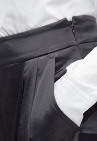 Μαύρη φούστα με πιετέ σχεδιασμό, που διαθέτει πλαϊνή τσέπη και ομαλή υφή υφάσματος. Μερικώς ορατό ένα λευκό πουκάμισο.