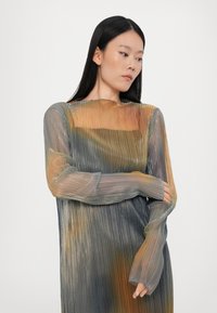 Durchsichtiges Kleid mit langen Ärmeln, das vertikale Falten in einem Farbverlauf von Blau, Grün, Orange und Gold aufweist. Es hat einen runden Ausschnitt und eng anliegende Ärmel.