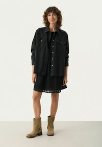 Schwarze Jeanshemd über einem schwarzen Spitzenkleid, kombiniert mit beigefarbenen Wildleder-Stiefeletten mit einem Riemenakzent. Einfaches, geschichtetes Outfit-Design.