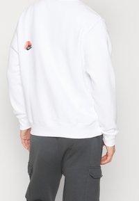 Vit sweatshirt med ribbade ärmslut och nederkant, med en liten färgglad Nike-logotyp på baksidan. Bärs tillsammans med mörkgrå cargobyxor.