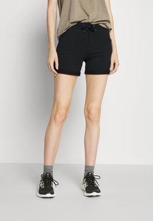 Person iført sorte shorts med snøre, grå strømper, sorte og hvide sportssko og en beige stribet skjorte, stående mod en ensfarvet baggrund.