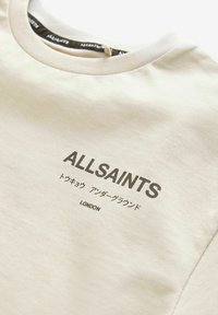 Krēmkrāsas T-krekls, kas izgatavots no mīksta kokvilnas, ar apaļu kakla izgriezumu, uz kura ir melni drukāti uzraksti: "ALLSAINTS," daudzvalodu rakstzīmes un "LONDONA."