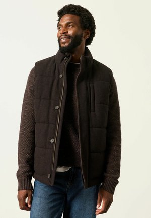 Mann lächelt, trägt eine dunkelbraune gesteppte Weste über einem strukturierten dunkelbraunen Pullover und blaue Jeans vor einem einfarbigen hellen Hintergrund.
