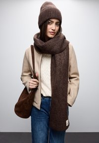 Brun strikket hue og tyk tørklæde, beige jakke over en cremefarvet sweater, mørke jeans og en brun semsket håndtaske, mod en neutral baggrund.