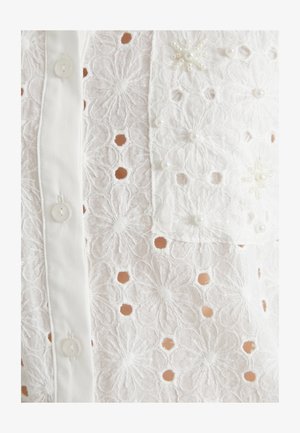 Weiße bestickte Bluse mit floralen Mustern, Cut-Out-Details und Perlenakzenten. Knopfverschluss vorne, halbtransparente Textur.