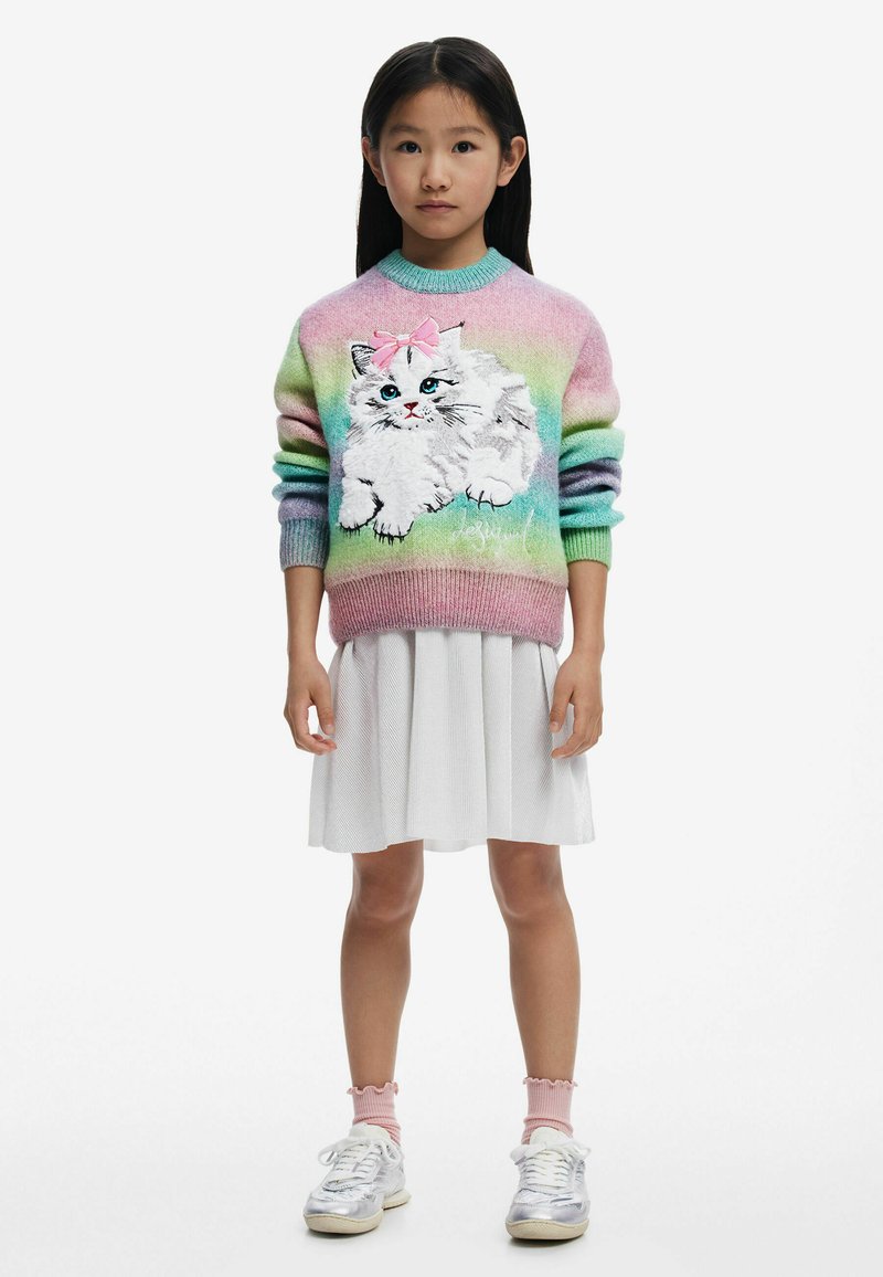 Bunt gestrickter Pullover mit einem Katzendesign und einer pinken Schleife, kombiniert mit einem weißen plissierten Rock und grauen Sneakers mit pinken Akzenten.
