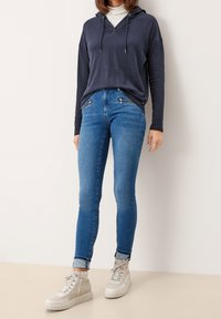 Tmavě modrá mikina s kapucí, světlé modré džíny s ozdobnými zipy a bílé vysoké tenisky s texturovanou podrážkou.