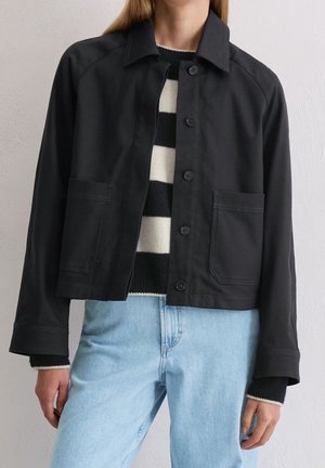 Henkilö käyttää mustaa napitettua takkia mustavalkoisen raidallisen villapaidan päällä, yhdistettynä vaaleansinisiiin farkkuihin.