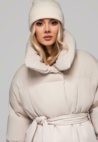Mladá žena s dlouhými blond vlasy, oblečená v krémovém pleteném beanie a oversized béžovém prošívaném kabátě s vysokým límcem, na jednotném pozadí.