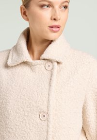 Abrigo de lana beige claro con superficie texturizada, que presenta un cuello ancho y dos grandes botones en la parte delantera para el cierre.