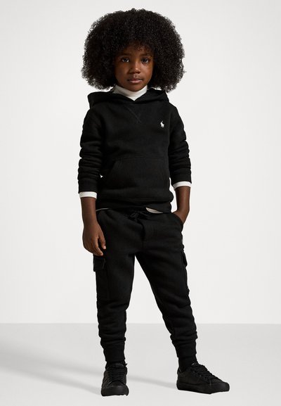 Svart fleece-hoodie med en framficka och vita detaljer, i kombination med svarta jogger-byxor med sidfickor och matchande svarta sneakers.