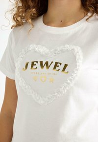 Vit bomullströja med volang i transparent kant i hjärtform. Har guldtexten: "JEWEL" och "SPARKLING OF JOY," tillsammans med grafiska ikoner.