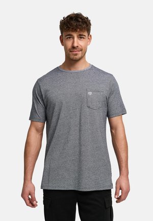 Jeune homme aux cheveux bouclés portant un t-shirt gris uni à manches courtes avec une petite poche poitrine et un pantalon noir.