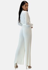 Monos de manga larga blancos con pliegues verticales, cintura ajustable y piernas anchas, complementados con sandalias de tacón alto beige.