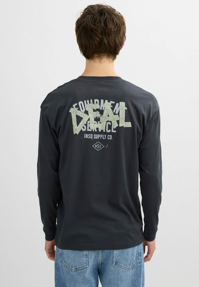 Camisa de manga larga de color gris oscuro que presenta un gran texto desgastado "DEAL" en la parte posterior, con gráficos blancos arriba y debajo. Ajuste casual.
