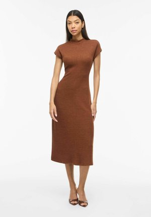 Femme portant une robe midi ajustée à manches courtes couleur rouille avec un col montant et des talons ouverts, debout devant un fond blanc.