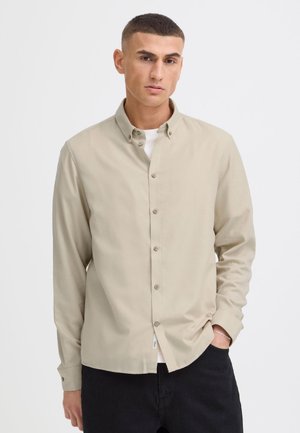 Jeune homme aux cheveux courts et foncés, portant une chemise beige à boutons sur un t-shirt blanc et un pantalon noir, debout avec une main dans la poche.