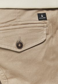 Bež pamučne hlače s preklopnom džepom, tamnim gumbom i svijetlosmeđim šavovima. Uključuju malu crnu oznaku s natpisom "JACK & JONES."