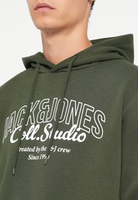 Žalia gobtuvė su raišteliu ir dideliu baltu užrašu "JACK&JONES Coll.Studio." Minkšta medžiaga su laisvu siluetu.