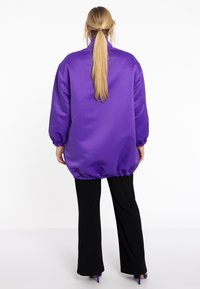 Veste oversize en satin violet avec des manches amples et un ourlet froncé, associée à un pantalon noir évasé et des chaussures à talons violettes.