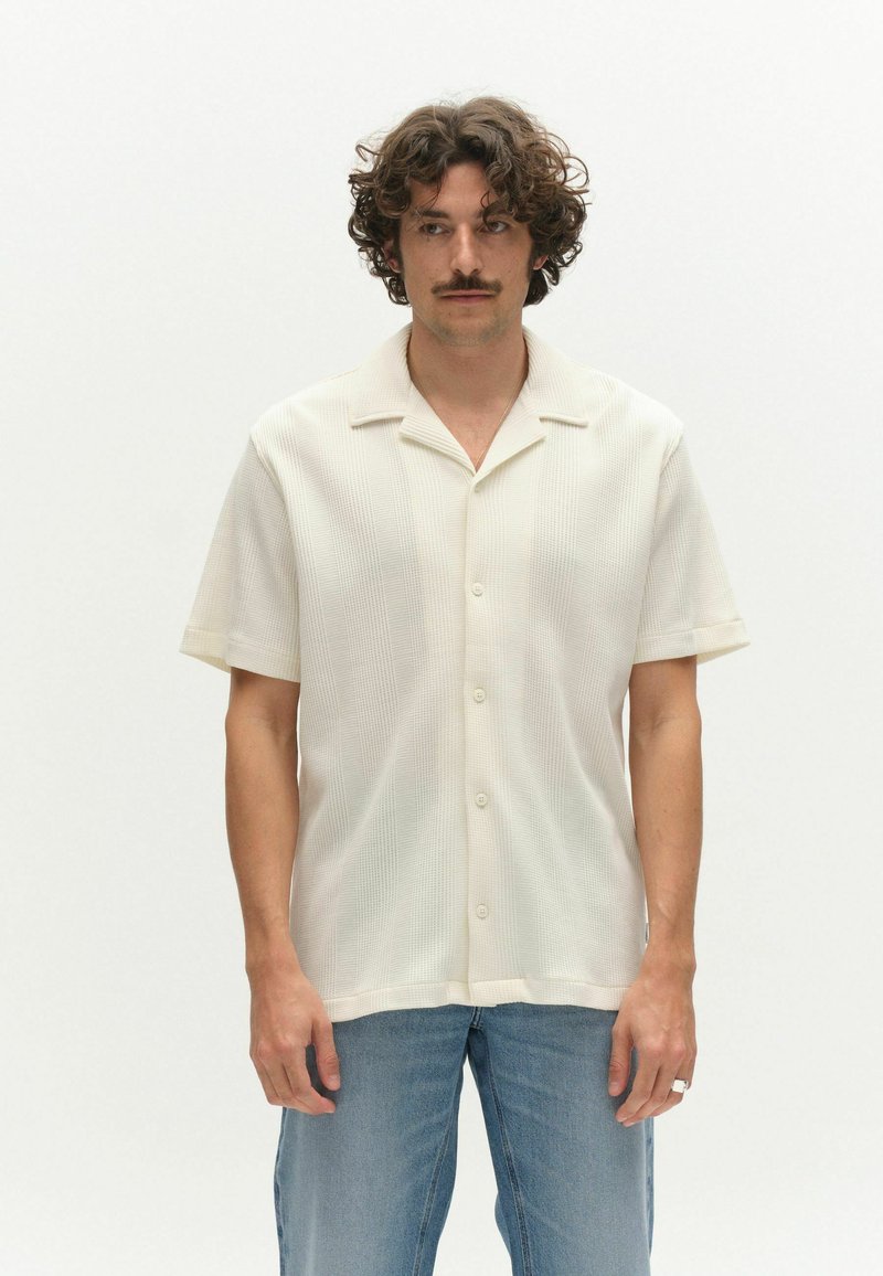 Göndör hajú és bajszos férfi rövid ujjú fehér, gombos ingben és világoskék farmernadrágban, egyszínű fehér háttér előtt áll.