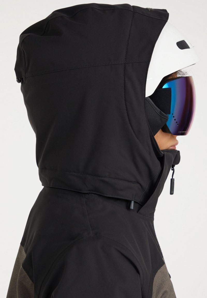 Schwarze kapuzenjacke mit einem kontrastierenden strukturierten grauen Paneel. Ausgestattet mit einem Reißverschluss und verstellbarer Kapuze. Getragen mit einem weißen Helm und Spiegelbrille.