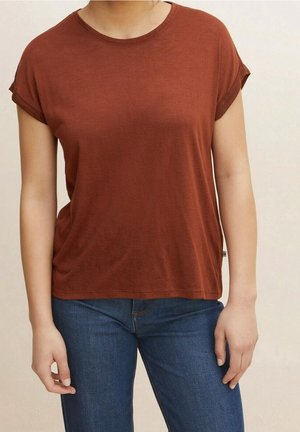 Kobieta ubrana w rdzawy, krótki rękaw koszulkę i niebieskie dżinsy, stojąca na jasnym, jednolitym tle.