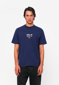 Navy blue cotton t-shirt with a round neckline and short sleeves. Features white text reading "USA SAIL GP TEAM" with a small flag graphic.