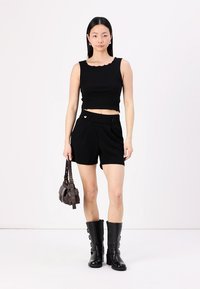 Top corto negro con canalé y bordes ondulados, pantalones cortos negros de tiro alto con detalle de botón y botas de cuero negro hasta la rodilla.