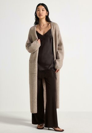 Kvinde iført en lang beige cardigan over en sort camisole med blonder og bukser med vide ben samt sorte åbne hæle.